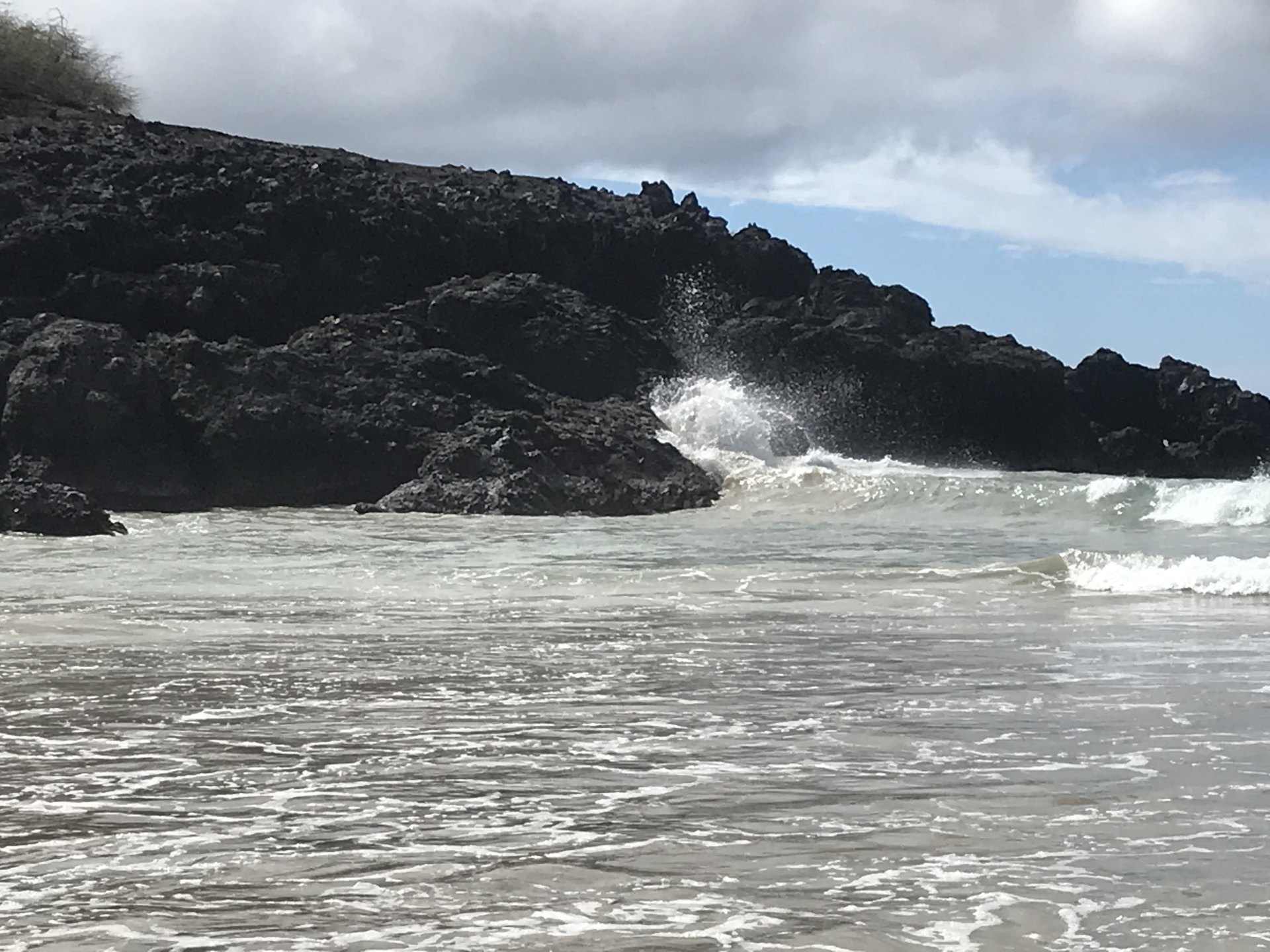 1 Corinthians 12 NASB.  Ocean waves crash against large black rock formations on the coast.