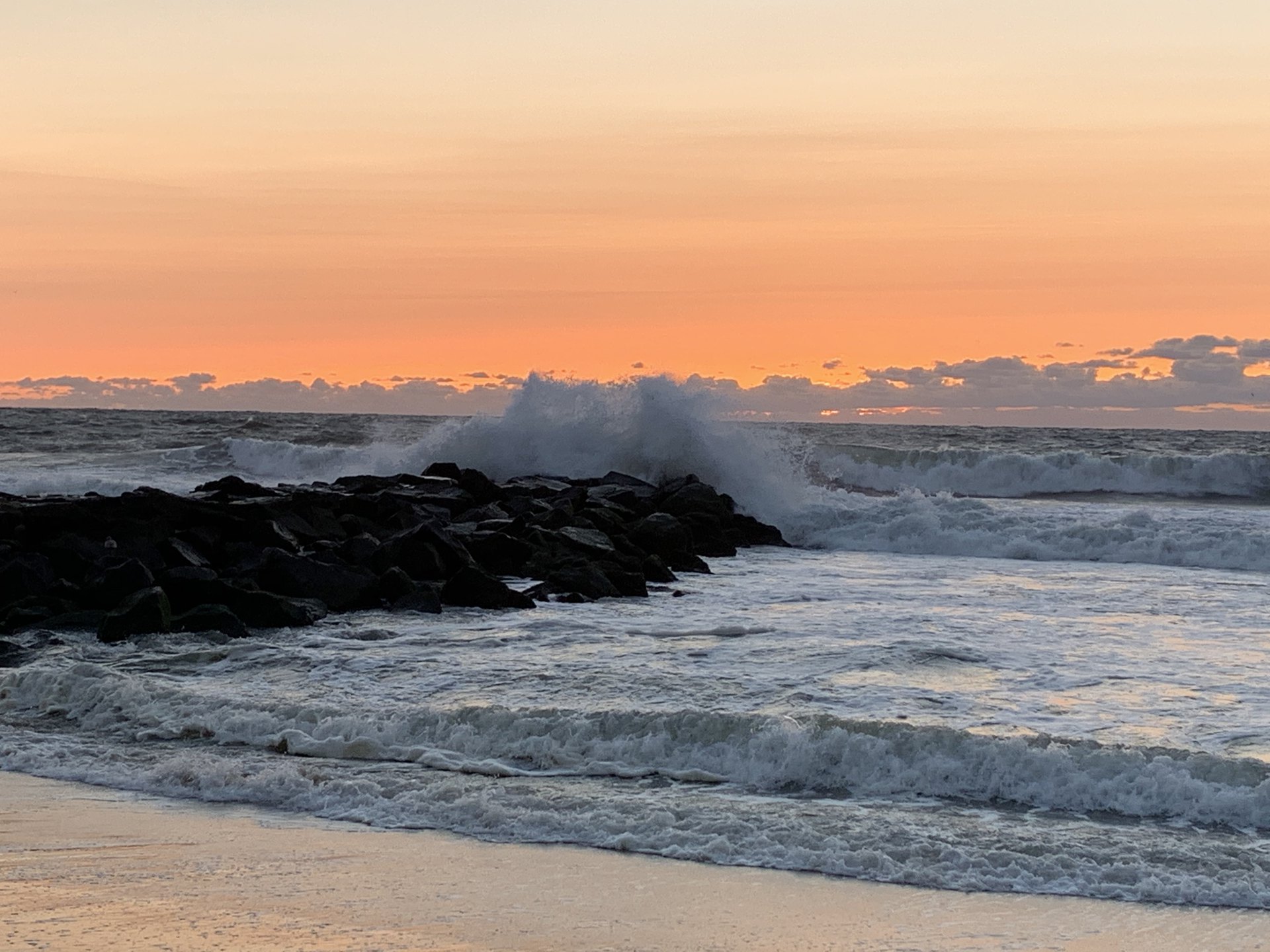 Acts 14 NASB.  Ocean waves crashing into the rocks against an orange backdrop sky.